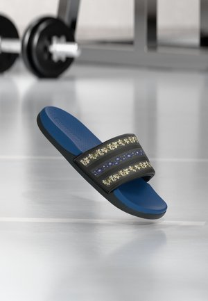 Sandale slide bleue avec sangle noire à motifs floraux, posée sur un sol réfléchissant avec des poids de gym flous en arrière-plan.
