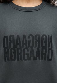 Σκούρο γκρι φούτερ με μεγάλα, ανάποδα μαύρα γράμματα που γράφουν "NØRGAARD" και "REGAARD" με έντονη, μοντέρνα γραμματοσειρά. Ομαλή υφή υφάσματος.