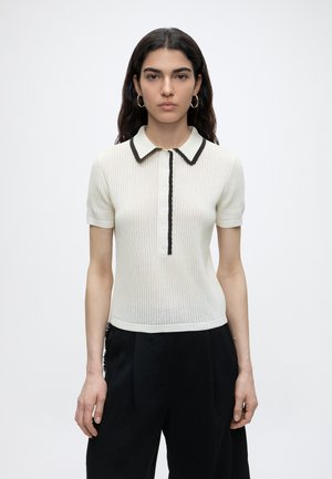 Młoda kobieta z długimi ciemnymi włosami nosi biały krótki rękaw sweterkowy polo z czarną lamówką i czarne spodnie, stojąc na jednolitym tle.