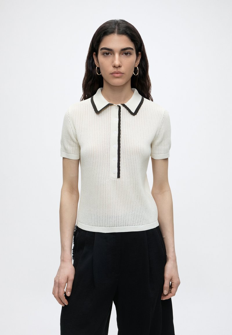 Νέα γυναίκα με μακριά σκούρα μαλλιά φοράει λευκή πλεκτή μπλούζα πόλο κοντομάνικη με μαύρο περιθώριο και μαύρο παντελόνι, στέκεται μπροστά σε απλό φόντο.