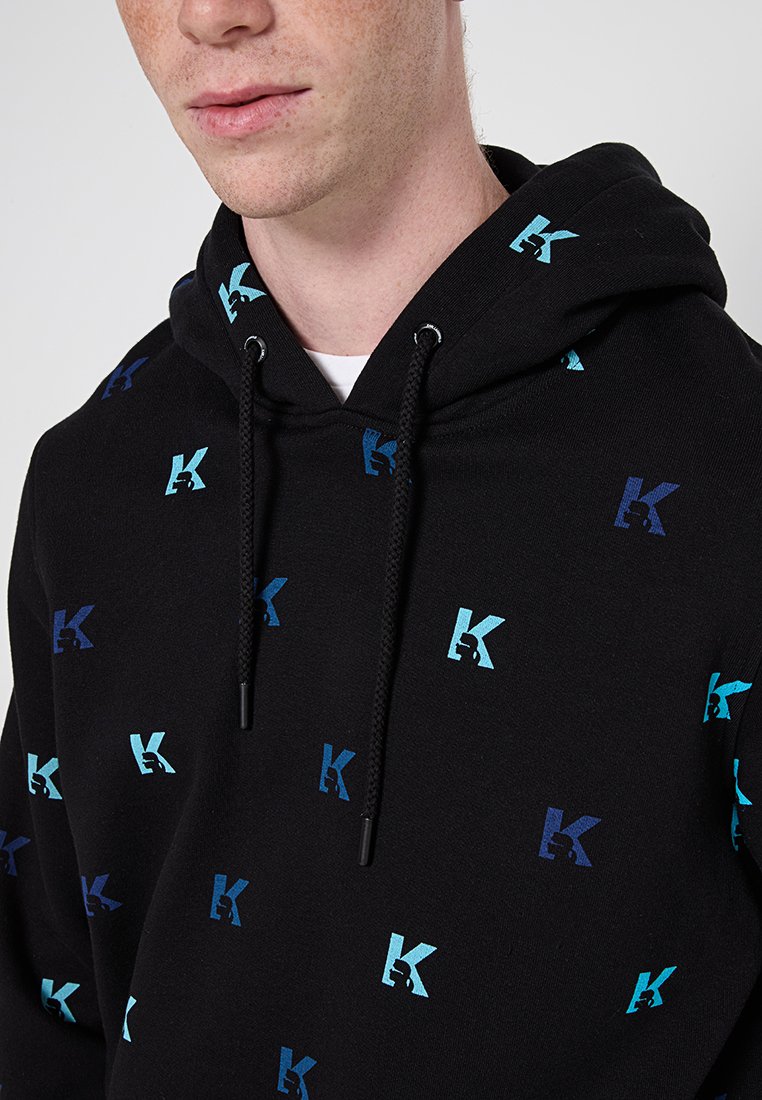 Sweat à capuche noir avec des logos "K" bleus et verts répétés, cordons de serrage et texture douce, idéal pour un style décontracté.