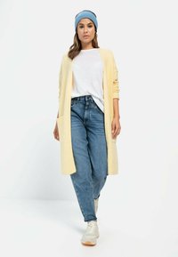 Lange, lichtgele cardigan met een open voorkant, gecombineerd met een witte blouse en hoge blauwe jeans. Witte sneakers en een blauwe gebreide beanie.