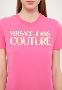 T-shirt en coton rose à manches courtes et col rond, avec un texte doré audacieux indiquant "VERSACE JEANS COUTURE" sur le devant.