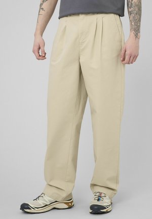 Homme portant un pantalon beige à jambes larges et des baskets beiges avec des bouts noirs, debout avec les bras tatoués visibles.