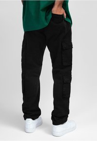 Pantalon cargo noir avec plusieurs poches latérales, fabriqué dans un tissu durable. Associé à des baskets blanches. Le haut n'est pas visible sur l'image.