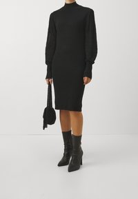 Abito nero a maglia con maniche lunghe a sbuffo strutturate e collo alto. Abbinato a stivali neri alti fino al ginocchio con tacco e una borsa rotonda all'uncinetto.