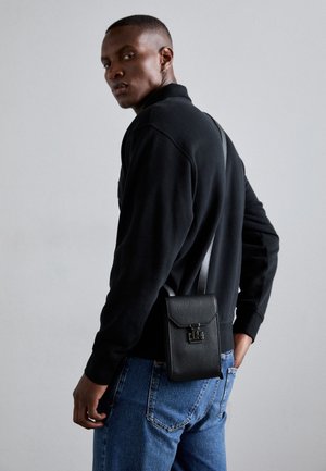 Mann trägt ein schwarzes Langarmshirt und blaue Jeans, trägt eine kleine schwarze Leder-Umhängetasche mit einem silbernen Verschluss.