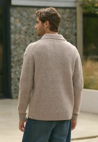 Beiger miteinander gestrickter Pullover mit umgeschlagenem Kragen, aus weichem Strickmaterial. Über blauen Jeans getragen, zeigt die Rückansicht.