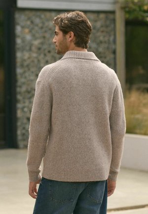 Beiger miteinander gestrickter Pullover mit umgeschlagenem Kragen, aus weichem Strickmaterial. Über blauen Jeans getragen, zeigt die Rückansicht.