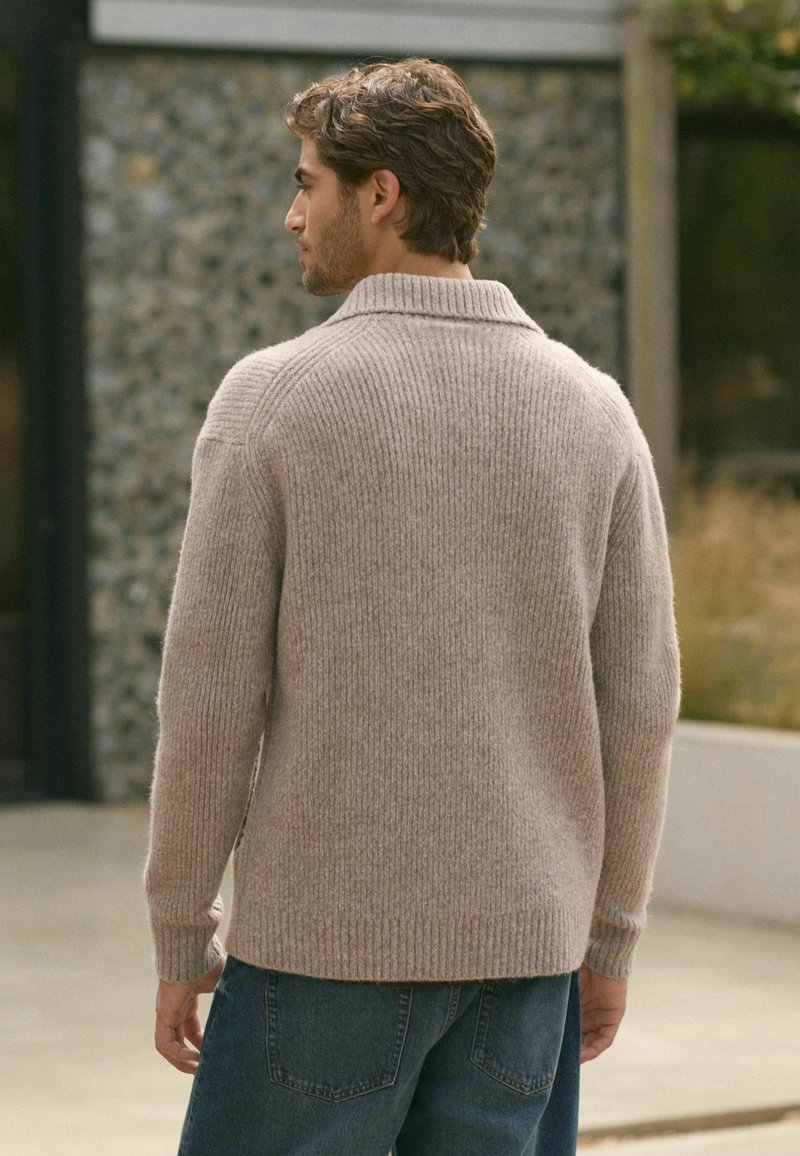 Beiger miteinander gestrickter Pullover mit umgeschlagenem Kragen, aus weichem Strickmaterial. Über blauen Jeans getragen, zeigt die Rückansicht.