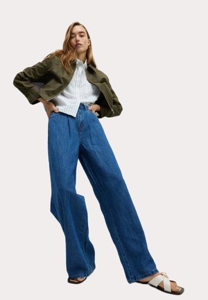 Chaqueta corta color oliva, camisa de botones blanca con rayas, vaqueros de corte ancho de mezclilla azul y sandalias negras con acentos blancos.