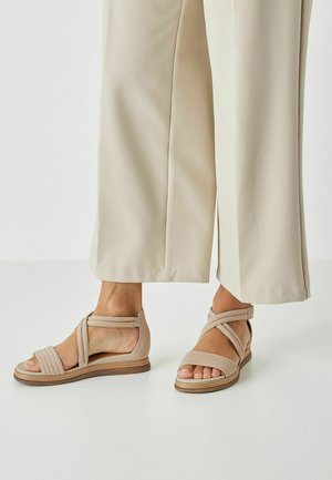 Beige sandaal met een gestructureerd bovenwerk, voorzien van kruisbandjes en een lage sleehak. Het ontwerp heeft een verstelbare enkelriem voor een goede pasvorm.