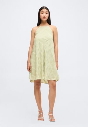 Donna in piedi, indossa un abito senza maniche verde chiaro al ginocchio con scollo a halter e sandali bianchi con cinturini e tacco alto.