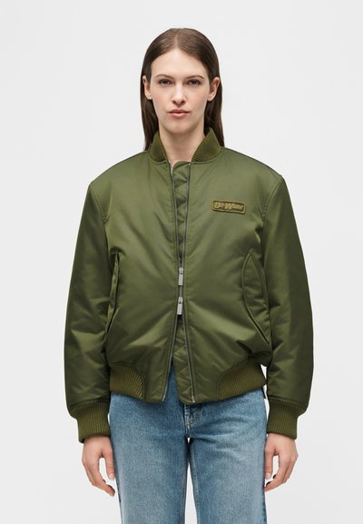 Γυναίκα με καστανά ίσια μαλλιά φοράει μια χακί μπουφάν τύπου bomber με δύο φερμουάρ, ένα πατς, και μπλε τζιν, σε ένα απλό φόντο.