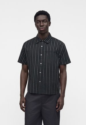 Jeune homme portant une chemise noire à manches courtes avec des rayures blanches verticales et un pantalon foncé, debout devant un fond blanc uni.