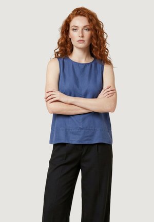 Młoda kobieta z rudymi kręconymi włosami, ubrana w niebieski top bez rękawów i czarne spodnie, stoi ze skrzyżowanymi ramionami na tle jednolitym.