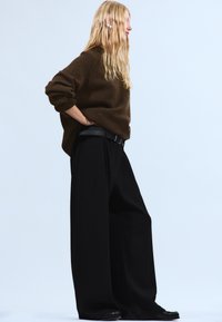 Mørkebrun strikket trøje kombineret med brede, sorte bukser og et sort bælte. Langt, glat hår, stående mod en lys baggrund.