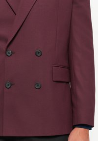 Dobbeltbrystet bordeaux blazer med sorte knapper, lommeklap og en mørkeblå ærmekant synlig på en persons arm.