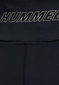 Svart atletiskt material med ordet "HUMMEL" i fet, ljust färgad text. Mjuk textur med flatlocksömmar synliga.