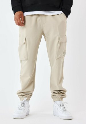 Pantalones cargo beige con dos bolsillos laterales, puños elásticos y una textura suave, combinados con zapatillas blancas. Sudadera negra visible en la parte superior.