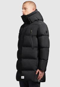 Svart puffjacka med hög krage, dragkedjeficka och texturerad quiltad design. Har en logotyp-patch nära sömmen.