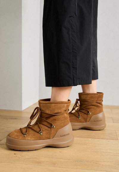 Bottes en suède marron avec un bout arrondi, un col moelleux, des lacets et une semelle texturée marron arborant un logo sur le côté. Associées à un pantalon noir court.