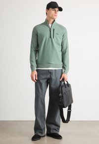 Grüner Pullover mit einem halbseitigen Reißverschluss, gerippten Bündchen und Logo. Weite Jeanshose und schwarze Accessoires, darunter eine strukturierte Tasche und eine Mütze.