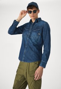 Sötétkék farmering, két zsebével a mellkasnál, gombos kialakítással és hosszú ujjakkal, olajzöld cargo nadrággal párosítva és napszemüveget tartva.