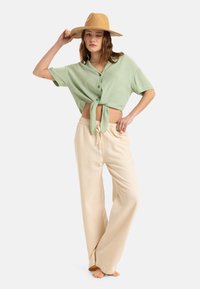 Camisa verde de manga corta y botones con un lazo en la cintura, combinada con pantalones anchos beige. Modelo lleva un sombrero de paja beige.