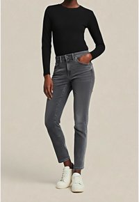 Personne portant un haut noir à manches longues, un jean slim gris avec la main dans la poche et des baskets blanches sur un fond uni.