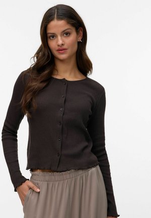 Kvinde med langt brunt hår iført en tætsiddende mørkebrun knapjakke og lysebeige højtaljede bukser, stående med den ene hånd i lommen.