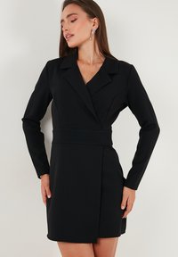 Woman wearing a fitted black long-sleeve blazer dress with a wrap front and wide collar, posing against a white background.