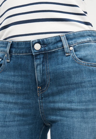 Μπλε τζιν παντελόνια με εφαρμοστό σχέδιο, με ασημένιο κουμπί, πορτοκαλί ραφές και μπροστινές τσέπες. Μπλούζα με ρίγες, μερικώς ορατή πάνω από αυτά.