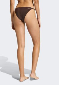 Hnědé bikiny s nízkým pasem vyrobené z texturované tkaniny, nabízející minimální zakrytí. Jemný detail loga na zadní straně.