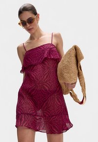 Fuchsiové průsvitné šaty s volánkovým lemem a zebrovým vzorem, s tenkými ramínky. Modelka drží pletenou hnědou tašku a nosí velké sluneční brýle.