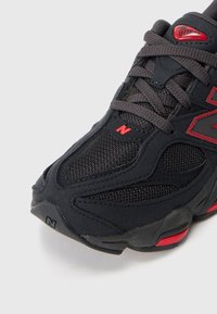 Shoe atlético preto com texturas de malha e camurça, apresentando detalhes em vermelho, biqueira redonda e logótipo proeminente. Os atacadores são cinzentos e tecidas de forma compacta.