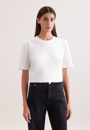 Kobieta o długich ciemnych włosach ubrana w prostą białą koszulkę z krótkim rękawem oraz czarne, wysokie dżinsy, stoi na jasnym tle.