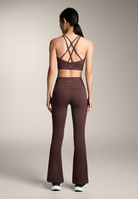 Brązowy stanik sportowy z krzyżującymi się ramiączkami z tyłu i pasującymi leginsami z rozkloszowanym dołem, charakteryzującymi się gładką fakturą i wysokim stanem. Widoczne białe sneakersy.