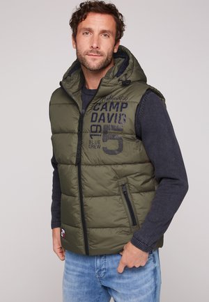 Mann trägt grüne gepolsterte ärmellose Jacke mit Kapuze, dunkelblauen Pullover und blaue Jeans, steht vor hellem einfarbigen Hintergrund.