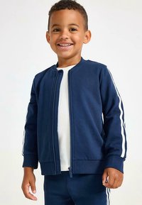 Marineblauw jack met rits en witte strepen langs de mouwen, geribbelde manchetten en zoom. Draagt een wit shirt eronder, onderdeel van een bijpassend trainingspak.