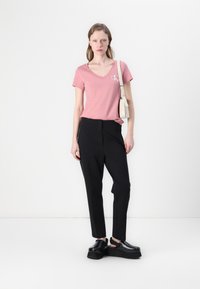 Roze V-hals t-shirt met een klein logo, zwarte op maat gemaakte broek en chunky zwarte schoenen. Eenvoudige tas hangt van de schouder. Neutrale achtergrond.