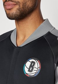 Black athletic top with a gray collar, featuring a circular logo with a stylized 'B', basketball design, and colorful accents in red, blue, and white.