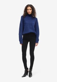 Tamsiai mėlynas megztinis su aukšta apykakle ir raštuota tekstūra, derinamas su juodomis liesomis džinsais ir atgal atvirais juodais kulkšnies batais.