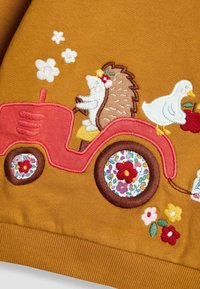 Hímzett pulóver vörös traktort ábrázol, mellette borz és kacsa található, mustár sárga háttér előtt, virágos részletekkel díszítve.