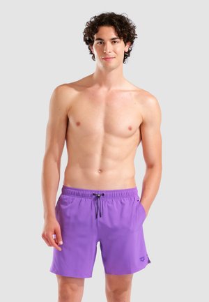 Joven con pelo rizado que lleva pantalones de baño morados con cordón, de pie con una mano en el bolsillo frente a un fondo liso.