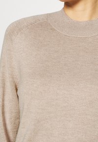 Beżowy sweter z dzianiny z ryżowym splotem, okrągłym dekoltem i wszytymi rękawami. Tkanina wydaje się miękka i elastyczna.