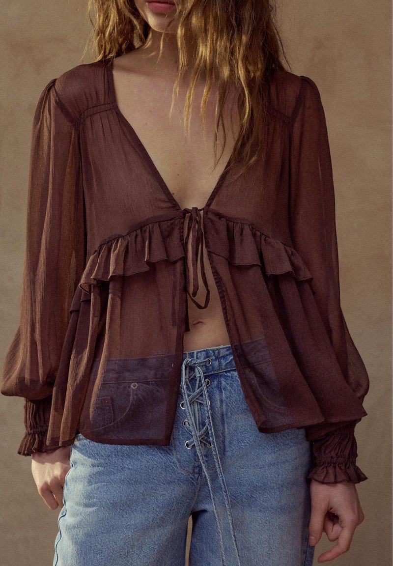 Femme portant un chemisier marron transparent avec de longues manches bouffantes et des volants, noué à l'avant, associé à un jean bleu taille haute à lacets.