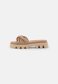 Sandale en caoutchouc beige avec une sangle nouée texturée, semelle plate présentant des stries superposées et un motif de traction crème contrasté.