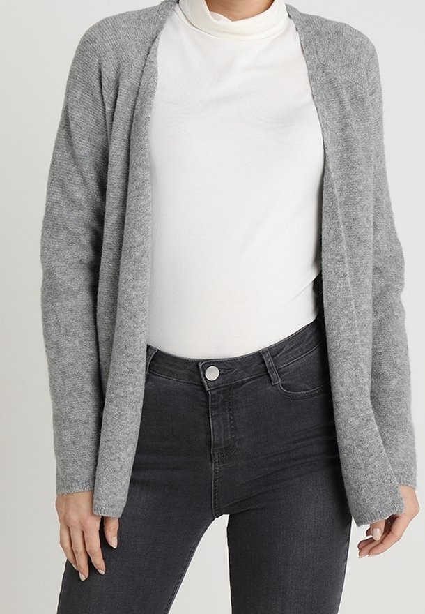 Personne portant un col roulé blanc, un cardigan gris ouvert sur le devant et un jean noir taille haute, debout les bras détendus le long du corps.