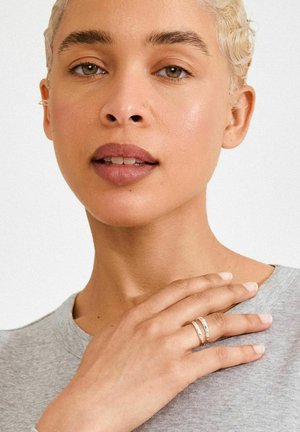 Sudraba gredzeni uz rokas, kas atrodas uz pelēkas krekla; fonā ir balts, izceļot sejas iezīmes un siltu ādas toni.